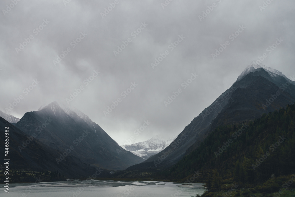 Atmospheric mountain landscape with gray mountain lake among dark rocks with snow under gray cloudy sky. Dramatic highland scenery with alpine lake in overcast weather. Gloomy mountain view in valley.