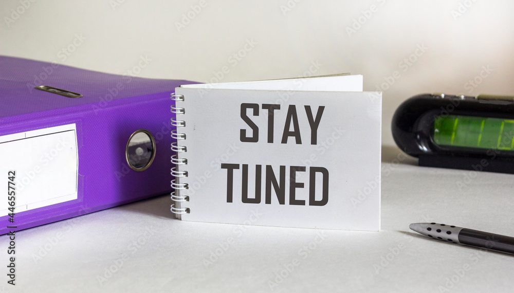 On a white background, a white notebook with the inscription STAY TUNED with a pen, a folder for documents and a pen with a clock On a white background, a white notebook with the inscription STAY TUNED with a pen, a folder for documents and a pen with a clock