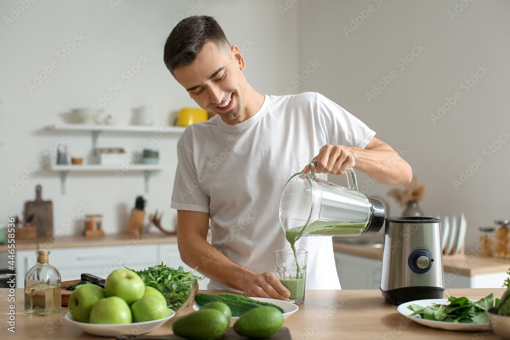 Fototapeta premium Young man pouring healthy green smoothie into glass in kitchen