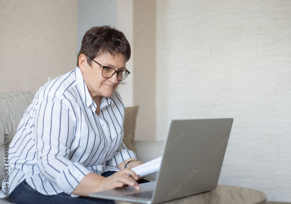 Portrait of mature lady in eyeglasses working on laptop at home office. Senior woman taking notes during online work meeting or webinar. Portrait of mature lady in eyeglasses working on laptop at home office. Senior woman taking notes during online work meeting or webinar.