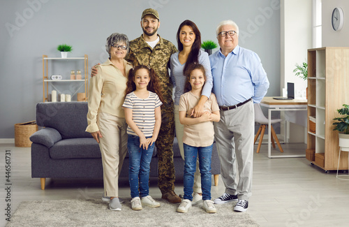 Soldier comes back home from military service. Indoor family portrait of grandma, grandpa, mom, children, and happy veteran dad in camo uniform standing in living-room, looking at camera and smiling