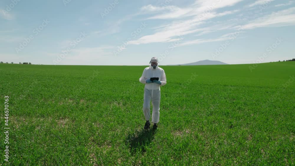 A worker, a biologist in a protective suit with a mask on his face, walks through a farmland, wind power plants in the background. A worker, a biologist in a protective suit with a mask on his face, walks through a farmland, wind power plants in the background.
