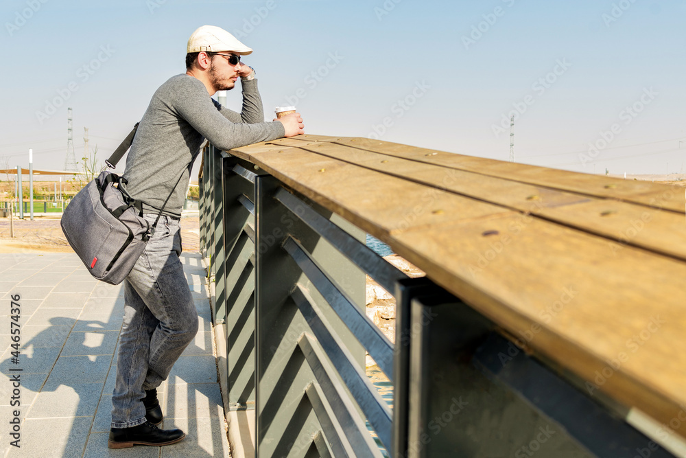Side view of relaxed man, drinking coffee, waiting on a bridge, on a sunny spring day.