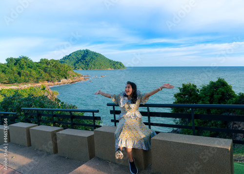 Asian woman tourist in dress sit and feel fresh with happy arm raised at seat beside walkway on Noen Nang Phaya viewpoint with blue sea and mountain view, travel attraction at Chanthaburi, Thailand photo