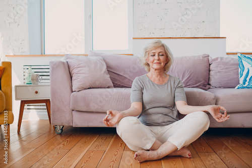 Senior adult smiling woman practicing yoga at home living room. Elderly relaxed female sitting in lotus pose and meditating at home