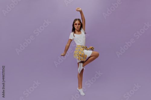Charming curly girl in white t-shirt and skirt moves on isolated purple background. Stylish teenager has fun in studio, rises arm and leg.