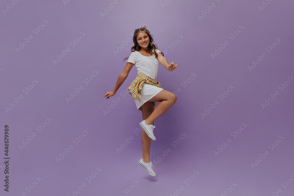 Tanned teenager jumping on isolated. Curly happy young girl in white dress and checkered shirt moves on purple background.