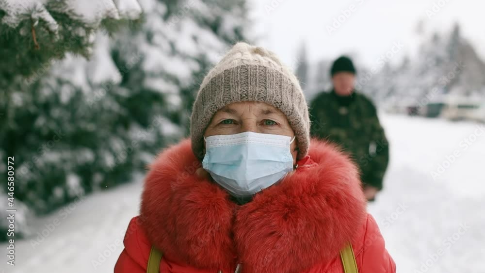 Adult senior woman in protective medical mask looking at the camera in the winter park. Social distancing during Coronavirus Covid 19 pandemic. Beautiful spruce trees branches sprinkled with white Adult senior woman in protective medical mask looking at the camera in the winter park. Social distancing during Coronavirus Covid 19 pandemic. Beautiful spruce trees branches sprinkled with white