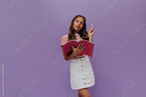 Charming young girl holds pen and pink notebook. Teenager has great idea and studies on purple background.