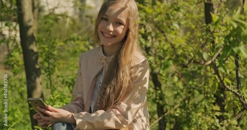 Portrait Smiling preteen girl using mobile phone sitting on the fallen tree in the park and smiling at camera. Girl has long blond hair