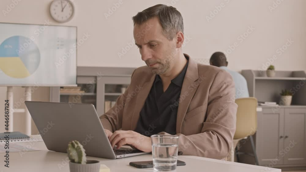 Waist-up of male office worker sitting at desk in office, typing on portable computer, then leaning back on chair, looking at results of his work proudly, smiling Waist-up of male office worker sitting at desk in office, typing on portable computer, then leaning back on chair, looking at results of his work proudly, smiling