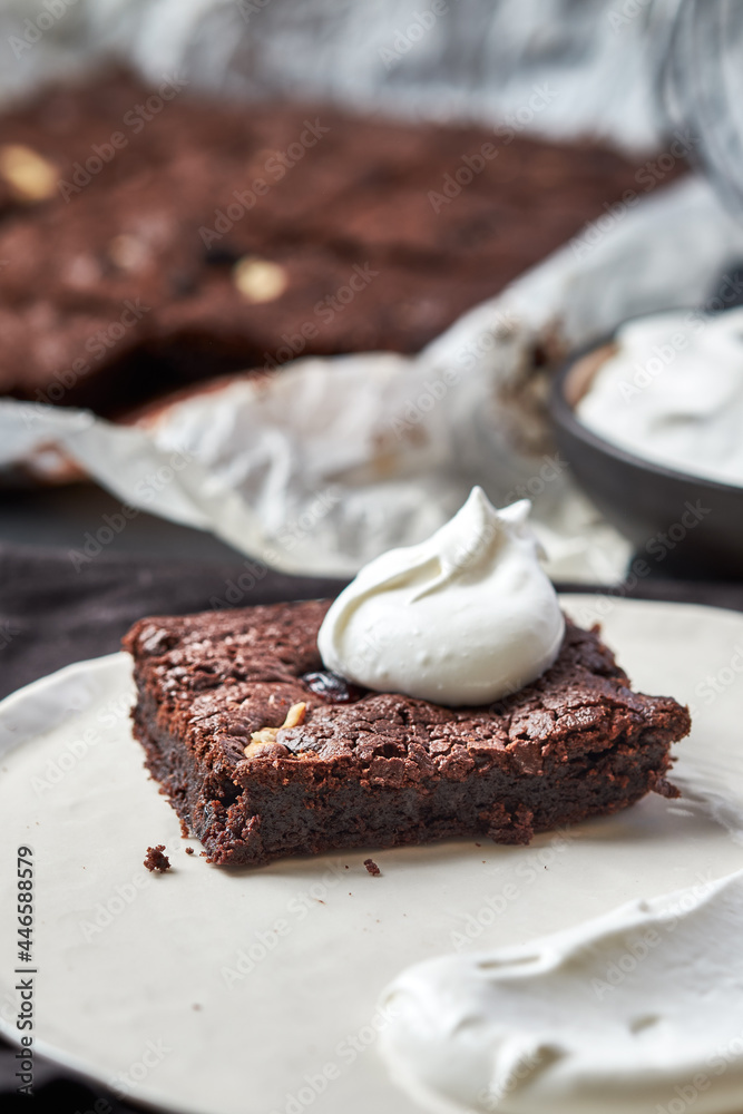 Brownie with ginger, dark chocolate coloured, on a white parchment lying on the gray table. A piece of brownie lies on a plate in the foreground. A bowl of cream stands near. Close up. 45 angle view Brownie with ginger, dark chocolate coloured, on a white parchment lying on the gray table. A piece of brownie lies on a plate in the foreground. A bowl of cream stands near. Close up. 45 angle view
