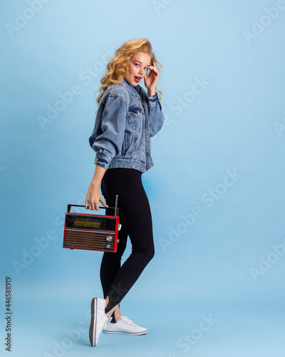 Portrait of pretty young girl in retro 90s fashion style, outfits posing isolated over blue studio background. Concept of eras comparison, beauty, fashion and youth. Side view