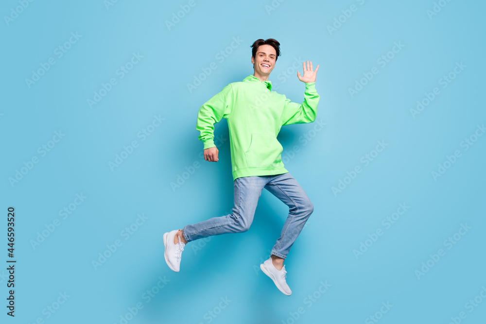Photo of sweet attractive young gentleman wear green sweatshirt smiling jumping high waving arm isolated blue color background Photo of sweet attractive young gentleman wear green sweatshirt smiling jumping high waving arm isolated blue color background