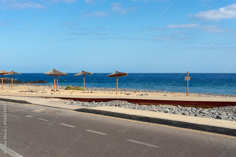 Solitary empty road passing through the small holiday resort known as San Blas and tranquil views towards a small artificial beach with white sand and straw parasols in Tenerife, Canary Islands, Spain