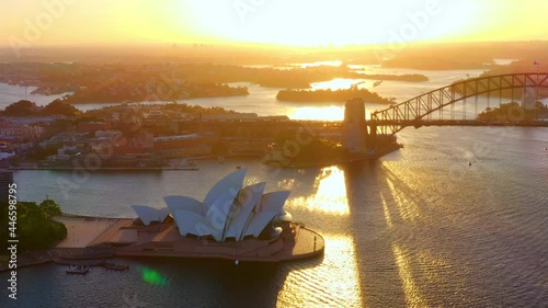 Golden Hour Over Sydney Harbour With Warm Sunlight On The Iconic Sydney Harbour Bridge And Opera House In Port Jackson Bay, NSW, Australia. aerial panning photo
