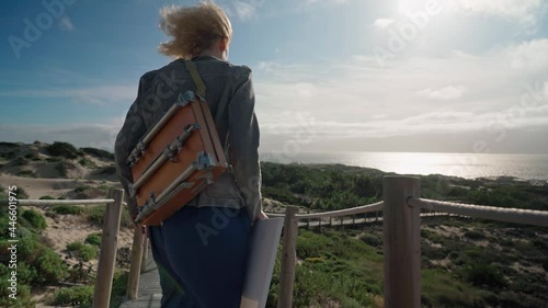 A middle-aged European woman artist with an easel on her shoulder walks towards the ocean.