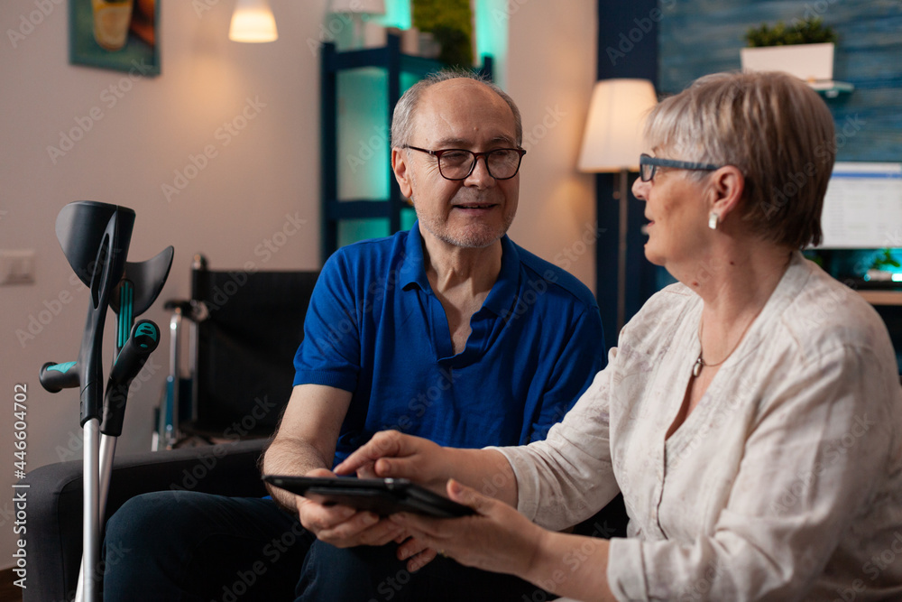 Obraz premium Elder couple using modern tablet with online internet on technology device in living room at home. Caucasian married man and woman with crutches and wheelchair for retirement support