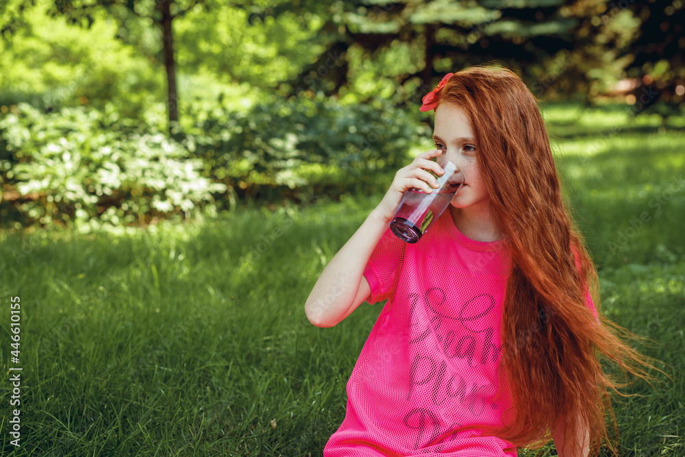 A beautiful young girl with red hair and in a red dress drinks clean water outdoors on a warm summer day. Healthy lifestyle concept. Space for text.