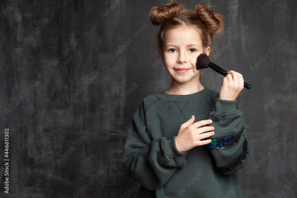 Fototapeta premium Beauty portrait of cute little girl model applying makeup with brush on background of blackboard with copy space. Pretty caucasian schoolgirl imitating putting powder and blush cosmetics on face.
