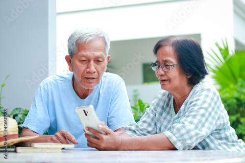 Asian elderly couple happy living at home Sitting in the garden holding a smartphone, chatting online with your child. Family concept, health care for the elderly in retirement age.