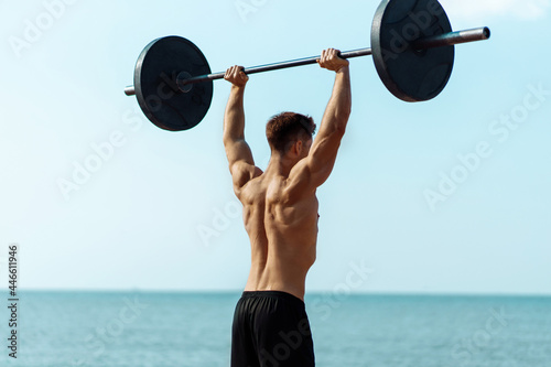 Muscular man training on the beach by the sea, doing exercises with a barbell for biceps, strong male press with a naked torso, sports concept