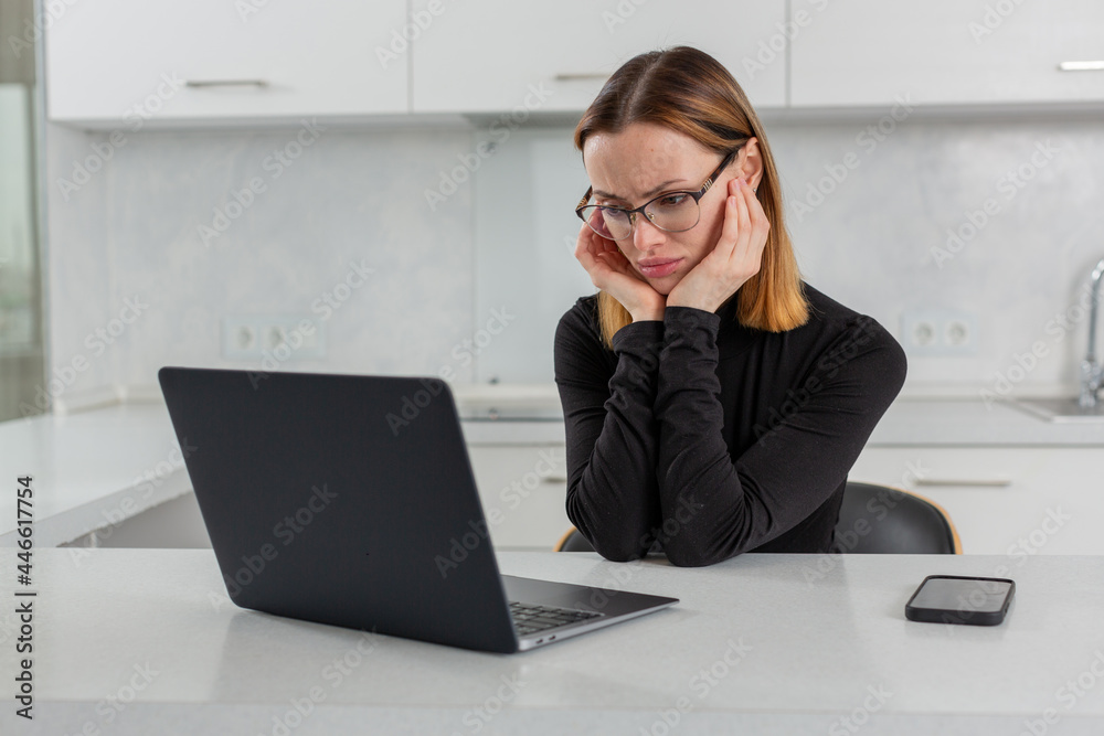 Girl in glasses sadly looks at the laptop monitor, sits at home in the white kitchen. High quality photo Girl in glasses sadly looks at the laptop monitor, sits at home in the white kitchen. High quality photo