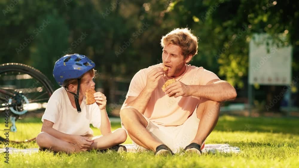 An attractive father and son are eating cookies while having picnic on the grass in the park at summer An attractive father and son are eating cookies while having picnic on the grass in the park at summer