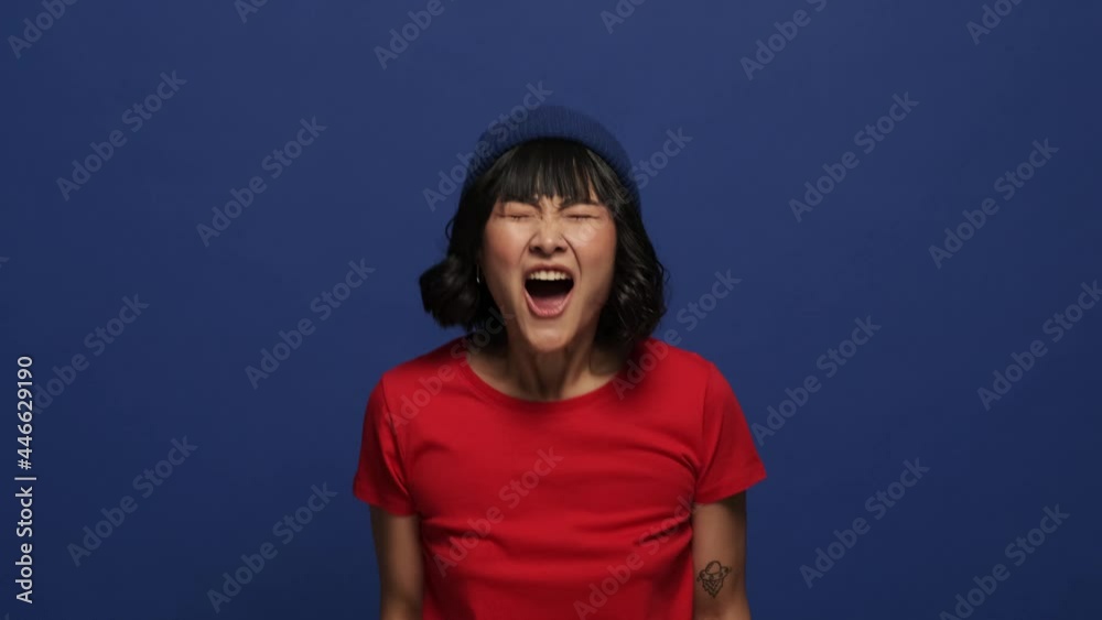 An angry emotional asian woman is screaming standing isolated over blue wall in the studio An angry emotional asian woman is screaming standing isolated over blue wall in the studio