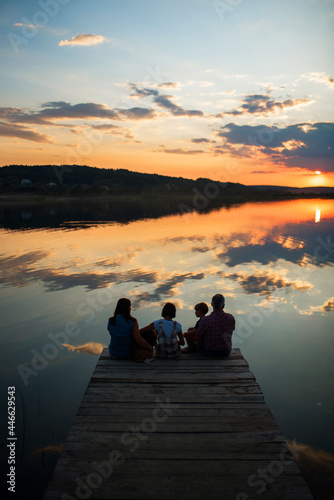 family of four people sitting on the shore on a wooden bridge of a large lake in summer and watching the beautiful sunset with dog back view