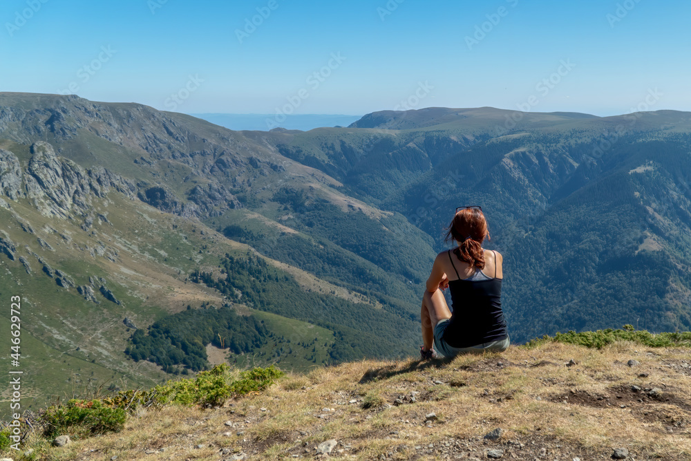 A woman sitting on top of a mountain in a relaxing mood and looking at a beautiful view of peaks, forests and blue skies
