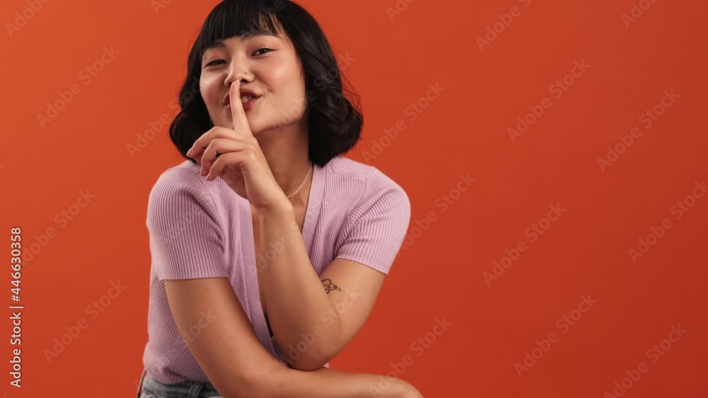 A beautiful asian woman is doing secret gesture to the camera isolated over orange wall in the studio A beautiful asian woman is doing secret gesture to the camera isolated over orange wall in the studio