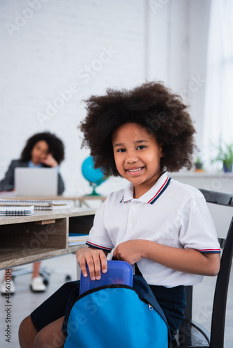 Happy african american pupil taking lunch box from backpack in classroom