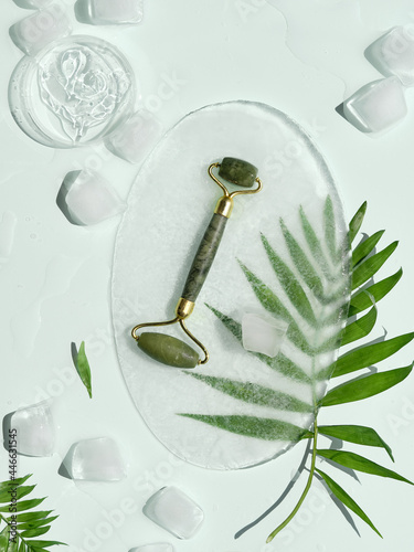 Self made moisturizer and green jade face roller on oval piece of ice and ice cubes. Exotic palm leaves on mint green background. Minimal flat lay, top view. Facial massage, handmade cosmetics.