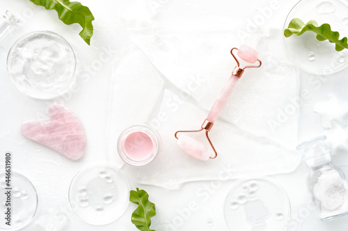 Fototapeta Naklejka Na Ścianę i Meble -  Pink quarts face roller, gua sha stone, pink cream in glass jar. Off white flat lay in green and pink with ice and exotic fern leaves. Beauty facial massage. Natural cosmetics laboratory.