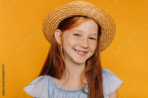 Happy excited redhead girl with freckles smiles sincerely on orange background. Long-haired child in boater and blue blouse looks into camera.