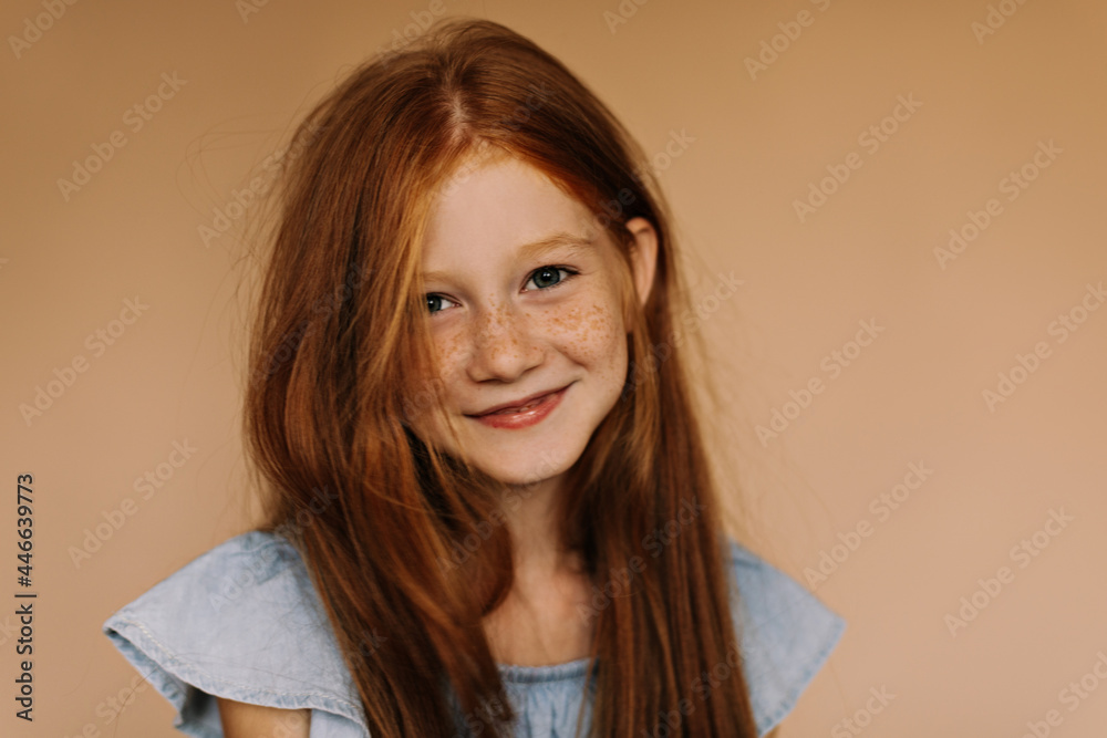 Positive girl with foxy long hairstyle and lovely freckles in modern blue clothes smiling and looking into camera on beige backdrop.. Positive girl with foxy long hairstyle and lovely freckles in modern blue clothes smiling and looking into camera on beige backdrop..