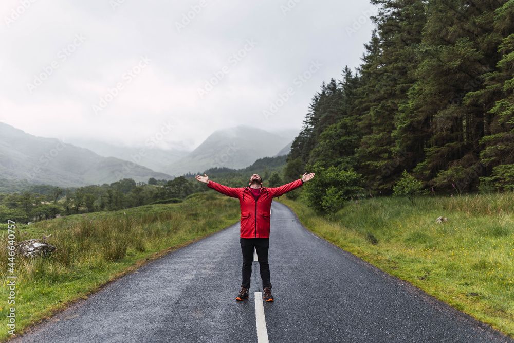 Traveler walking into the horizon on the lonely road in Scotland enjoying the scenery. He is wearing a red raincoat and surrounded by green trees and nature. Enjoying freedom