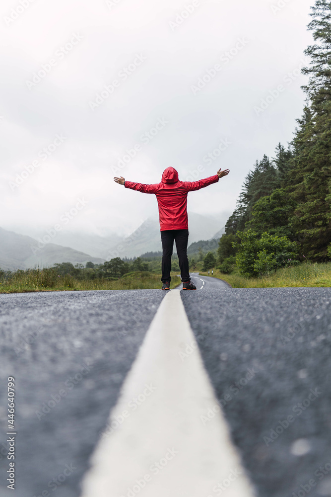 Traveler walking into the horizon on the lonely road in Scotland enjoying the scenery. He is wearing a red raincoat and surrounded by green trees and nature. Enjoying freedom