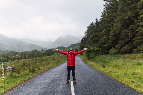 Obraz na płótnie Traveler walking into the horizon on the lonely road in Scotland enjoying the scenery. He is wearing a red raincoat and surrounded by green trees and nature. Enjoying freedom
w salonie
