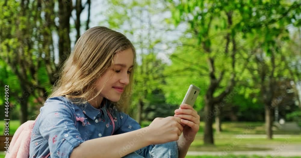 Cheerful school girl is online. She using mobile phone outdoors and smiling at camera. Preteen girl 10 years old sitting on the bench in the city park. Gimbal shot Cheerful school girl is online. She using mobile phone outdoors and smiling at camera. Preteen girl 10 years old sitting on the bench in the city park. Gimbal shot