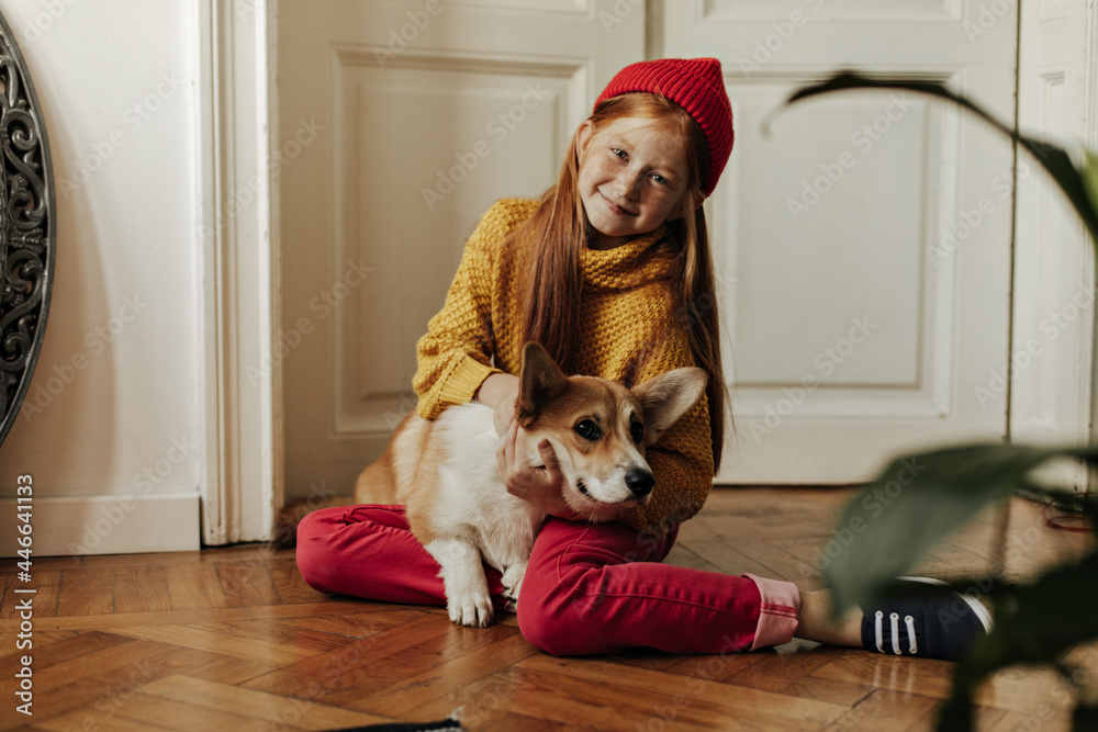 Joyful girl with ginger hair and cute freckles in trendy sweater, red pants  and bright cap smiling and petting corgi in apartment.. Stock Photo | Adobe  Stock