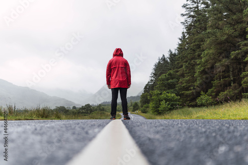 Obraz na płótnie Man in red raincoat walking in an empty road surrounded by a forest during a cloudy day in the Highlands, Scotland
w salonie