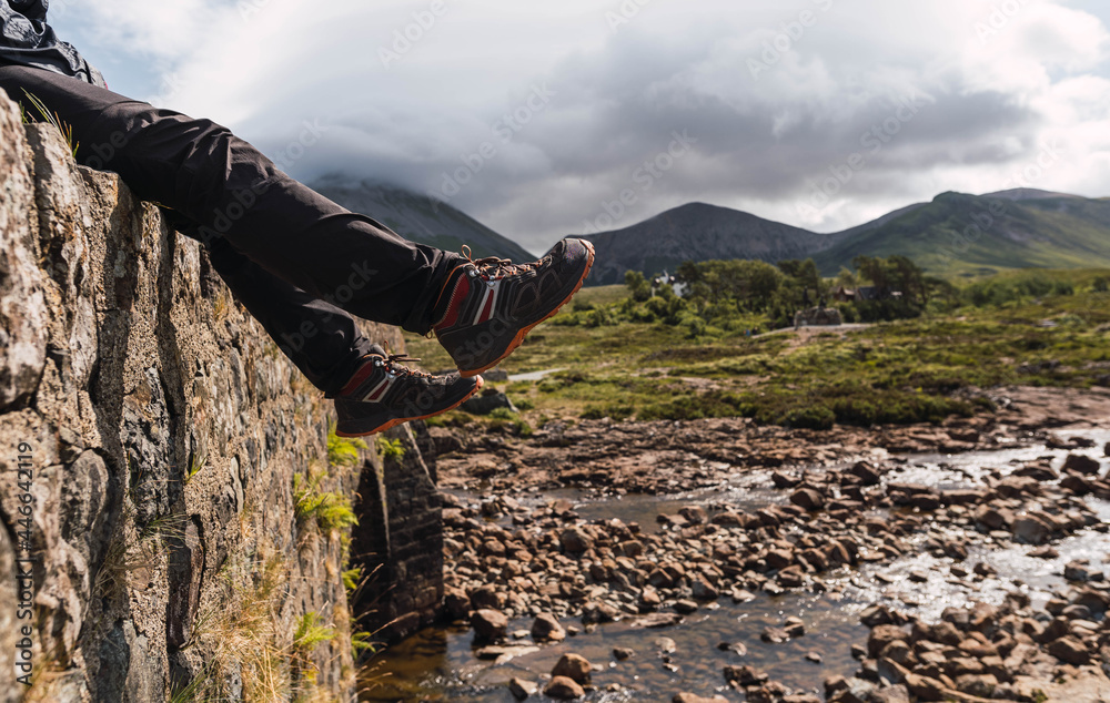 Young man sitting on the rock bridge while enjoying Scotland hill view at the Highlands, in Scotland. He is wearing hiking clothes. Sitting on an old vintage bridge in Sligachan, Isle of Skye