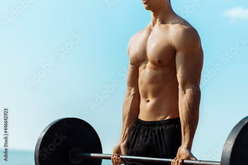 Muscular man training on the beach by the sea, doing exercises with a barbell for biceps, strong male press with a naked torso, sports concept