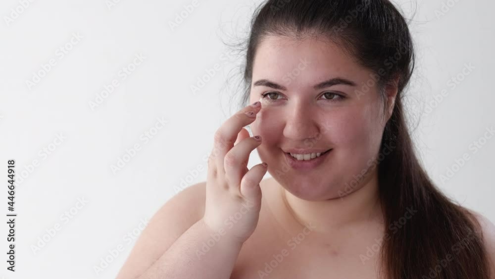 Skin moisturizing. Body positive. Facial treatment. Morning routine. Smiling overweight obese plus size woman in white towel applying under eye face cream after shower isolated on neutral. Skin moisturizing. Body positive. Facial treatment. Morning routine. Smiling overweight obese plus size woman in white towel applying under eye face cream after shower isolated on neutral.