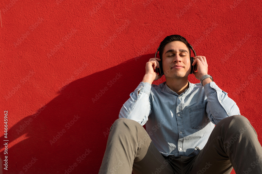 Portrait of young latin man with headphones enjoying listening to music with closed eyes sitting on floor. Red wall on background Portrait of young latin man with headphones enjoying listening to music with closed eyes sitting on floor. Red wall on background