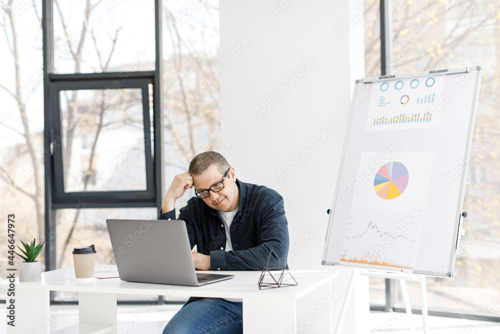 A man dressed in casual clothes works in the office with a laptop and expresses sad emotions.