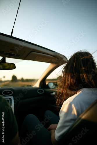 Rearview of an attractive brunette woman riding in the convertible car. Concept of summer adventures and new experiences. Strong sunlight during warm and long summer evenings.