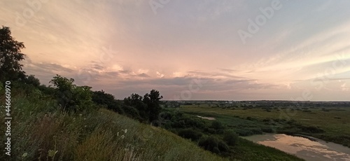 The evening sky is over the surface of the river. Hillside with silhouettes of trees. Bend of the river, stretching into the distance. The sun setting in the clouds. 